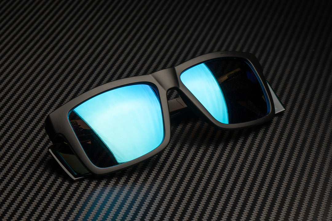 Laying on a table is the Heat Wave Visual Performance XL Vise Sunglasses with black frame, galaxy blue lenses and matching colored side shields. 