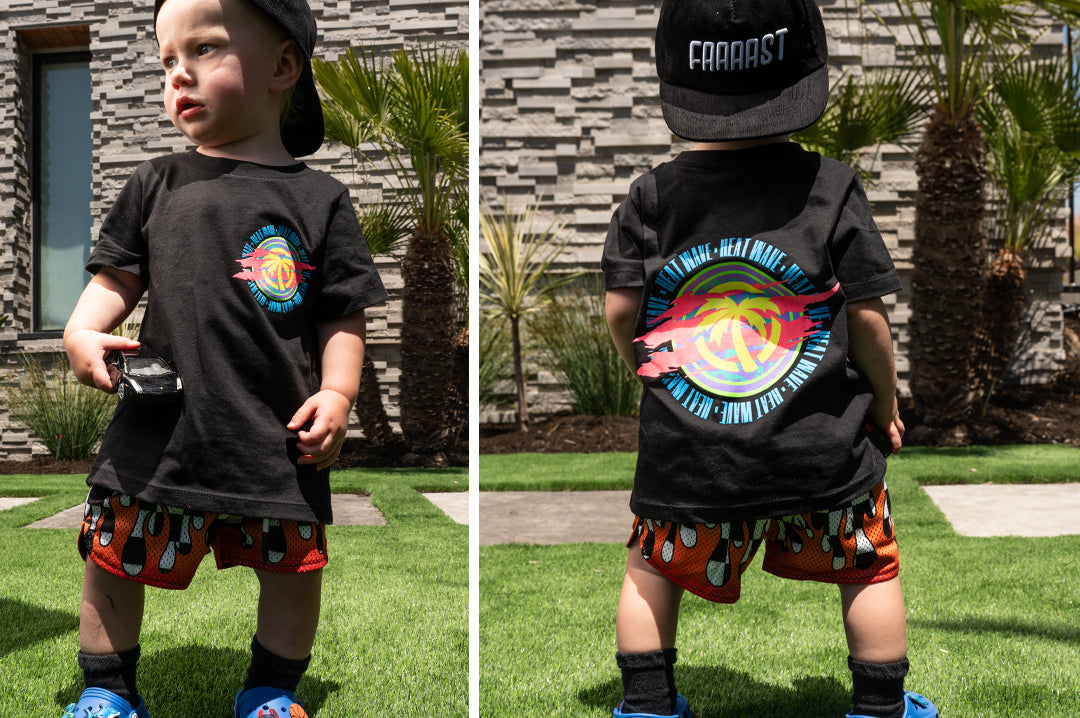 A toddler boy wearing the Heat Wave Saved by the Wave black t-shirt with backwards hat and swim shorts.